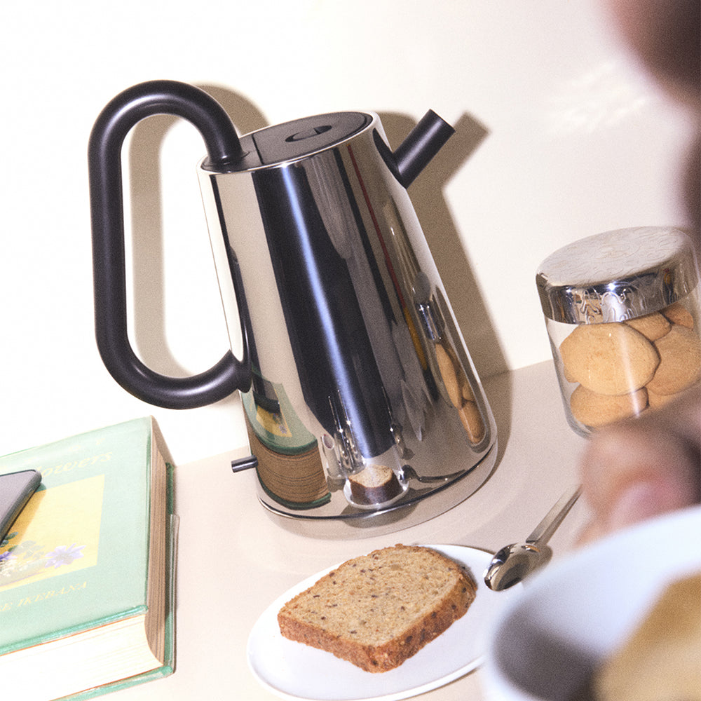 Shiny chrome kettle gleams beside toast, cookies, and a book—calm morning ritual of simple, tactile comforts.