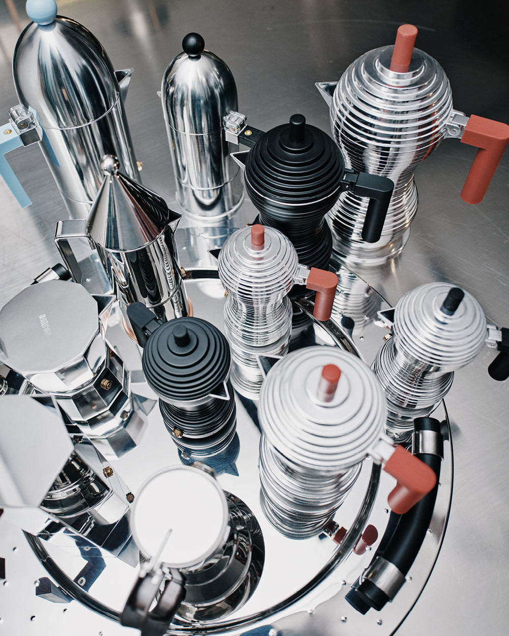 Sleek Alessi coffee makers in polished metal and bold colors, arranged artfully on reflective surface, embodying modern design elegance.