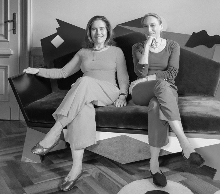 Two women sit confidently on a modern sofa, smiling warmly in a stylish, artistic setting.