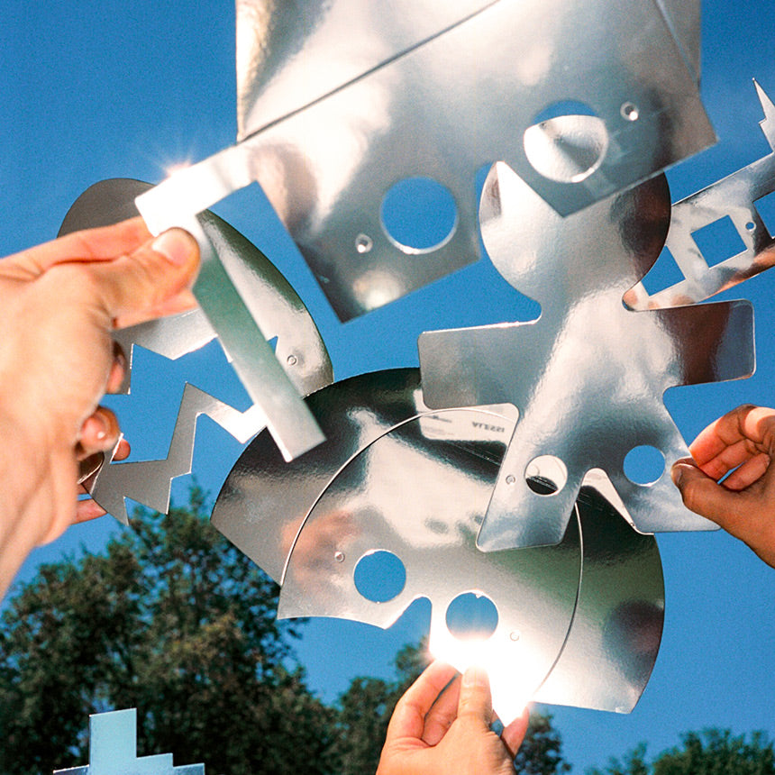 Hands assembling sleek, reflective metal puzzle pieces against a bright blue sky, showcasing Alessi MDW24’s artistic, modular design.