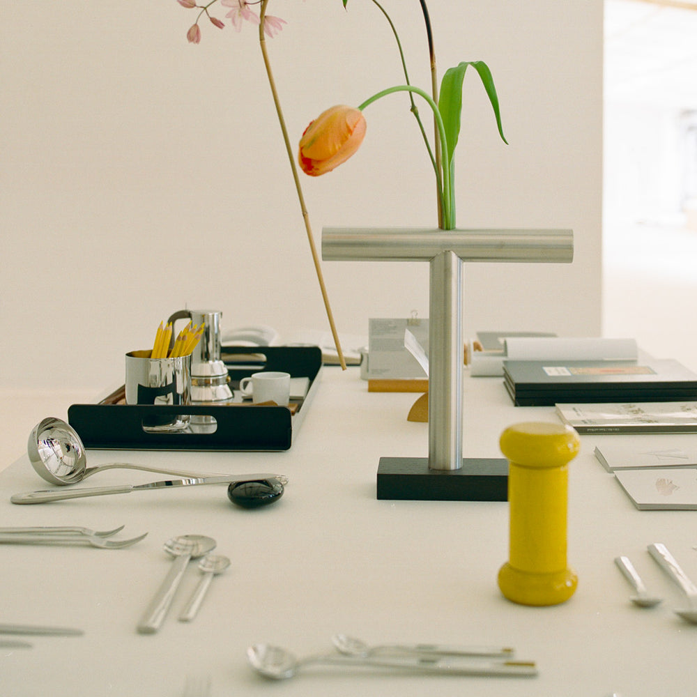 Minimalist table scene with sleek silverware, a yellow pepper mill, and a modern T-shaped vase holding delicate flowers, evoking calm and design elegance.