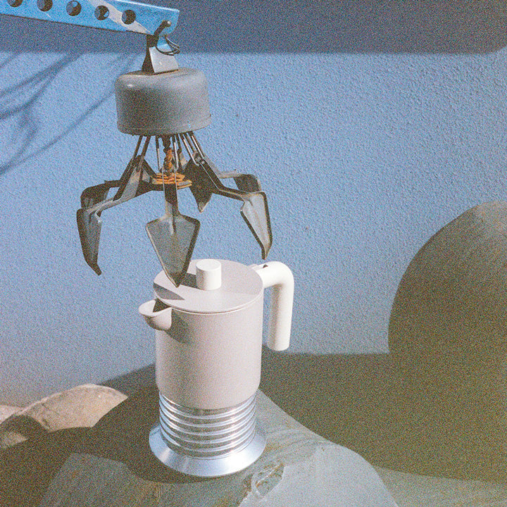 Minimalist white kettle with metallic base, held by robotic claw against cool blue backdrop, showcasing Philippe Malouin’s futuristic design.