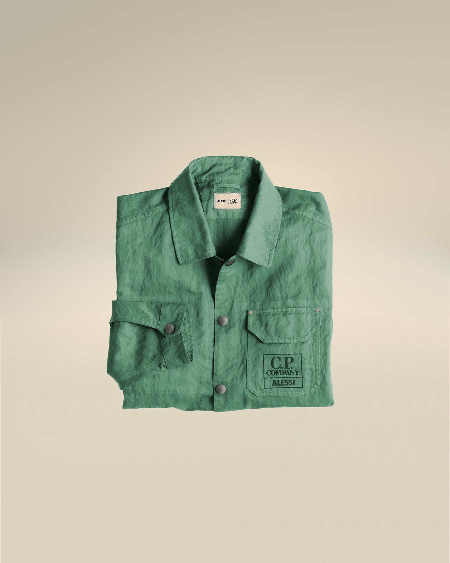 Green utility jacket by Alessi with a crinkled texture, featuring a C.P. Company ALESSI logo on the chest pocket, folded flat.