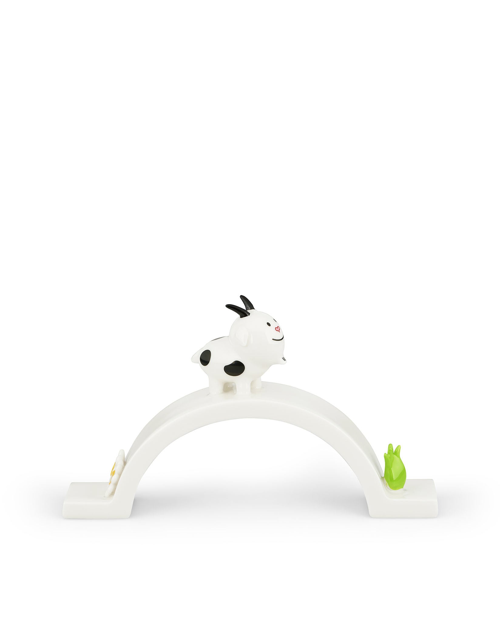 White goat ornamental figurine with black spots by Alessi, standing on a white arch base decorated with a yellow flower and green sprout.