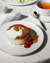 White ceramic Itsumo table set by Alessi, featuring a round plate, cup, saucer, and linen napkin on a white tablecloth.