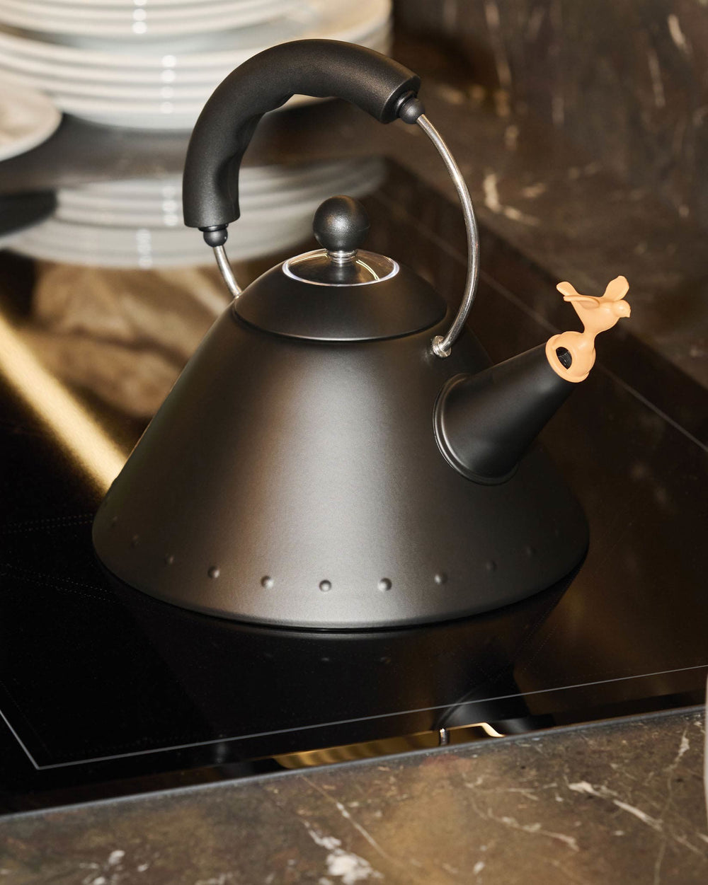 Matte black conical kettle by Alessi with a bird-shaped whistle and studded base, on an induction cooktop.