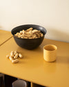 A large black bowl by Alessi, full of peanuts, displayed on a mustard yellow table with a yellow cup.