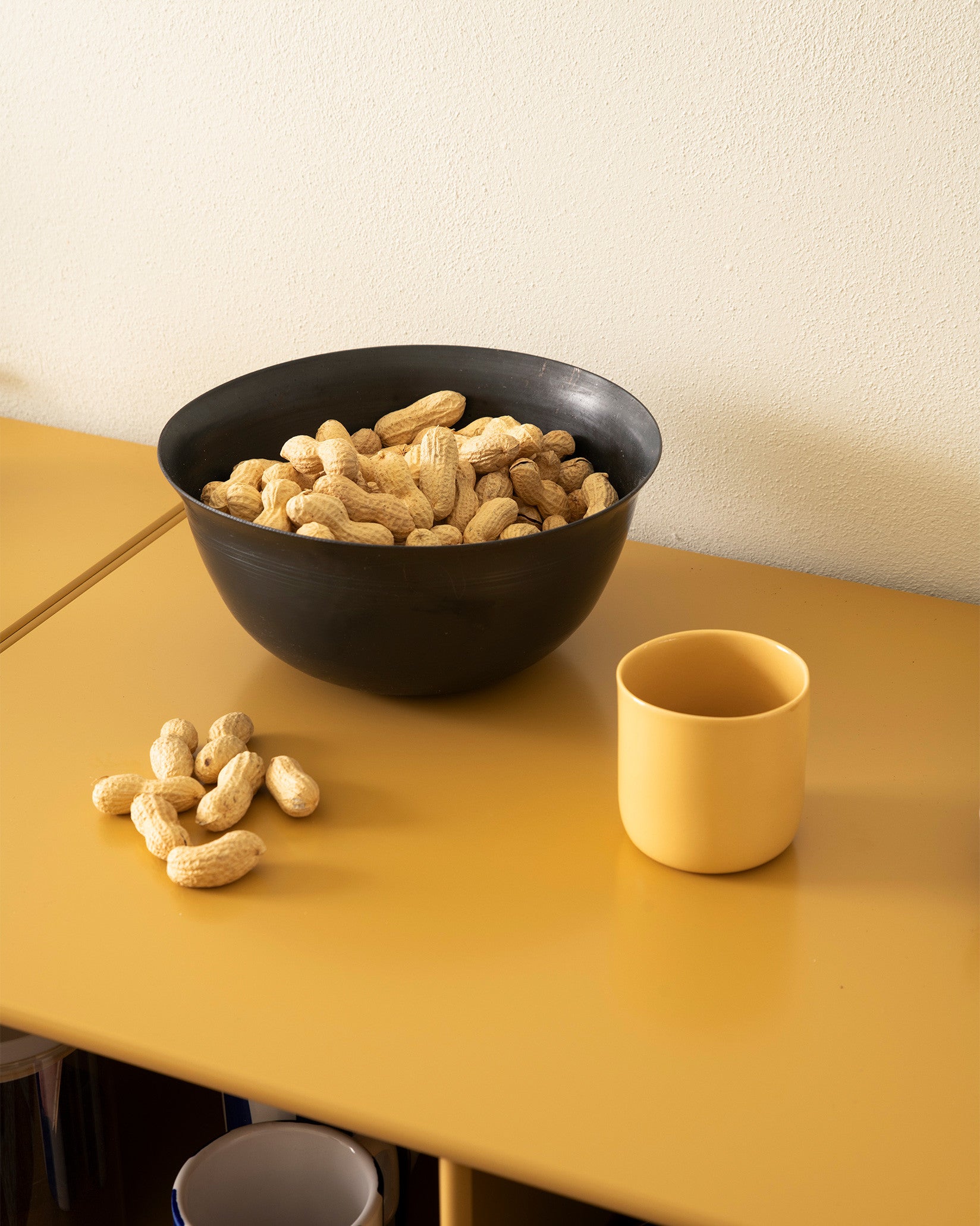 A large black bowl by Alessi, full of peanuts, displayed on a mustard yellow table with a yellow cup.