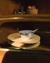 Silver metallic fragrance diffuser by Alessi with a lid and dark reed sticks, resting on two round marble trays.