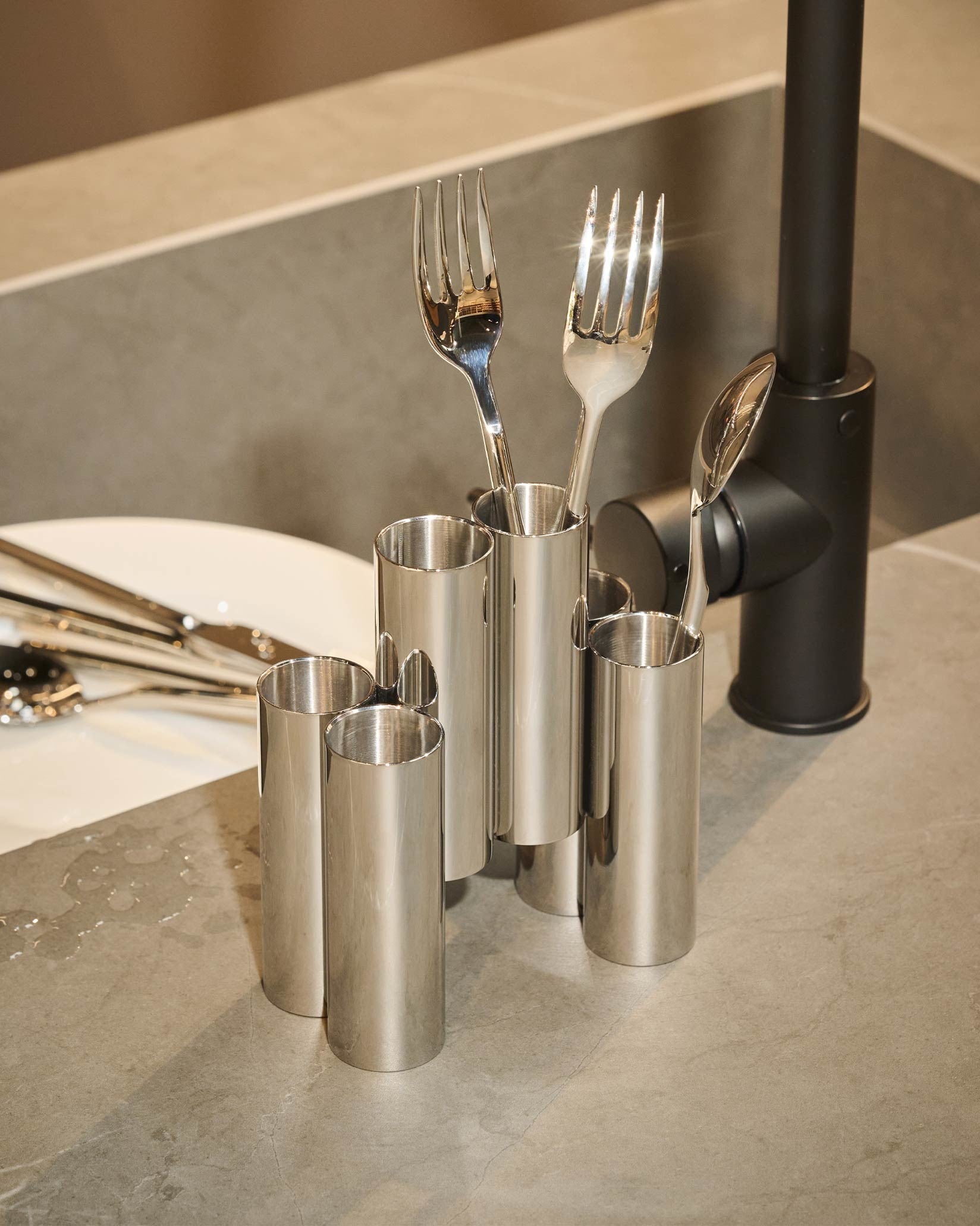 Interconnected cylindrical stainless steel cutlery holders by Alessi, holding silver forks and spoons on a wet gray counter.