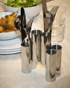 Polished stainless steel cutlery holders by Alessi, featuring multiple cylindrical vessels holding assorted flatware on a dining table.
