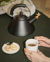 Black matte kettle by Alessi with a bird-shaped whistle, alongside cookies and a cup of tea.