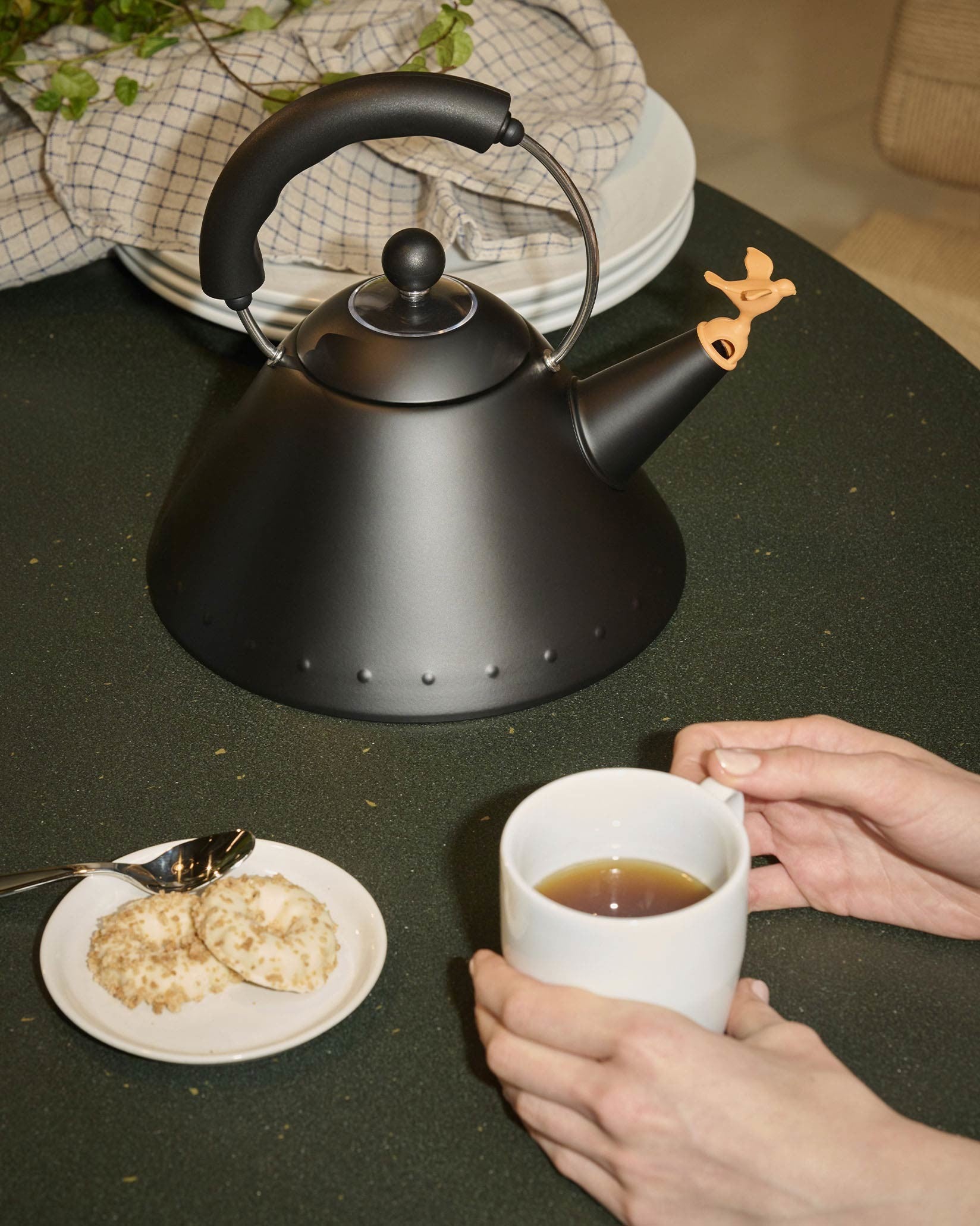 Black matte kettle by Alessi with a bird-shaped whistle, alongside cookies and a cup of tea.