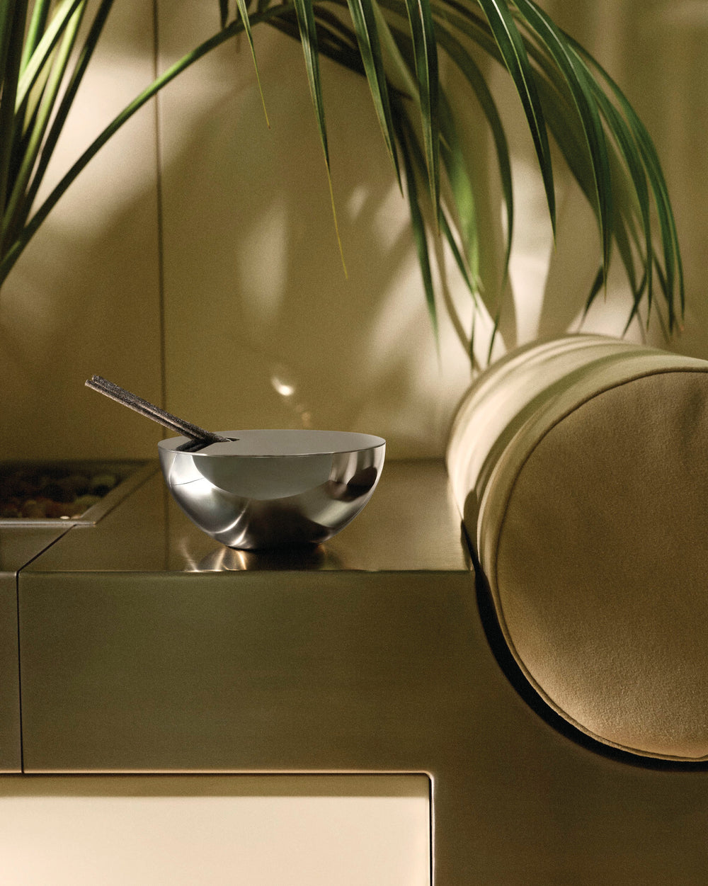 Silver polished metal bowl-shaped fragrance diffuser by Alessi, holding two dark incense sticks on a bronze surface with a green plant.