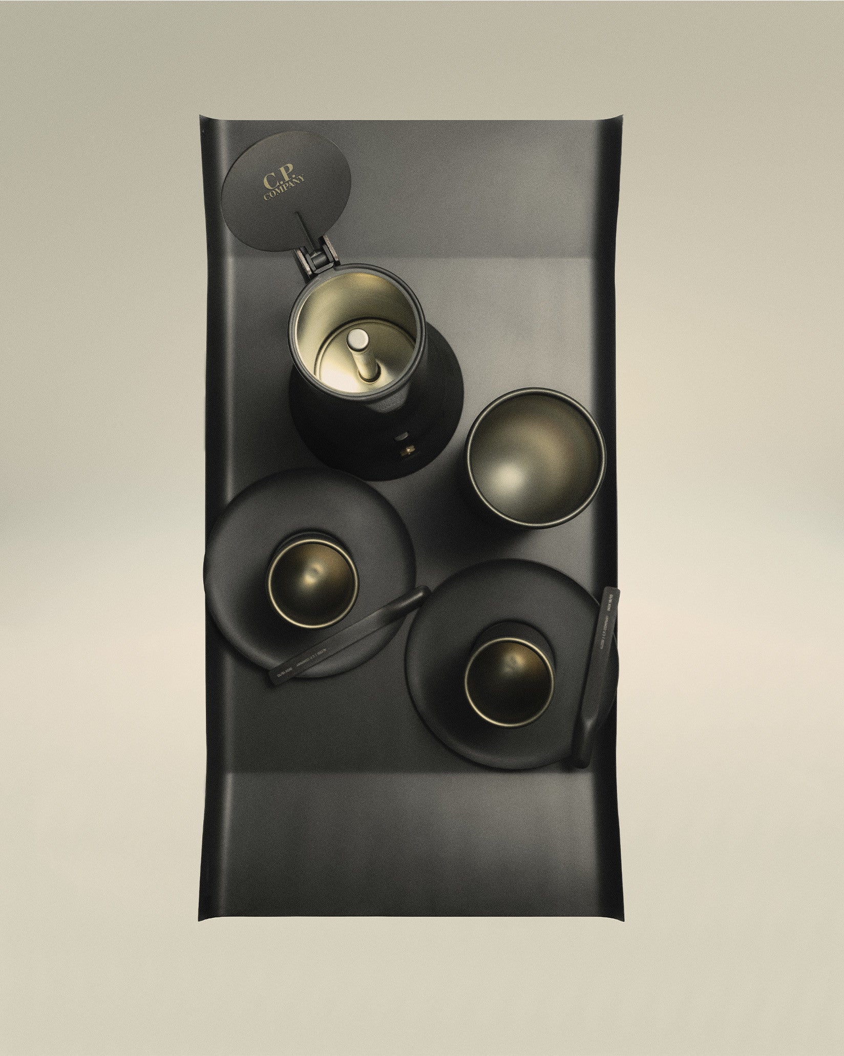 Dark grey metallic tray by Alessi filled with a black coffee maker, three cups, and spoons, all with golden interiors.