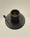Matte black cylindrical cup by Alessi with a gold interior rim, on a matching saucer and branded stirrer.