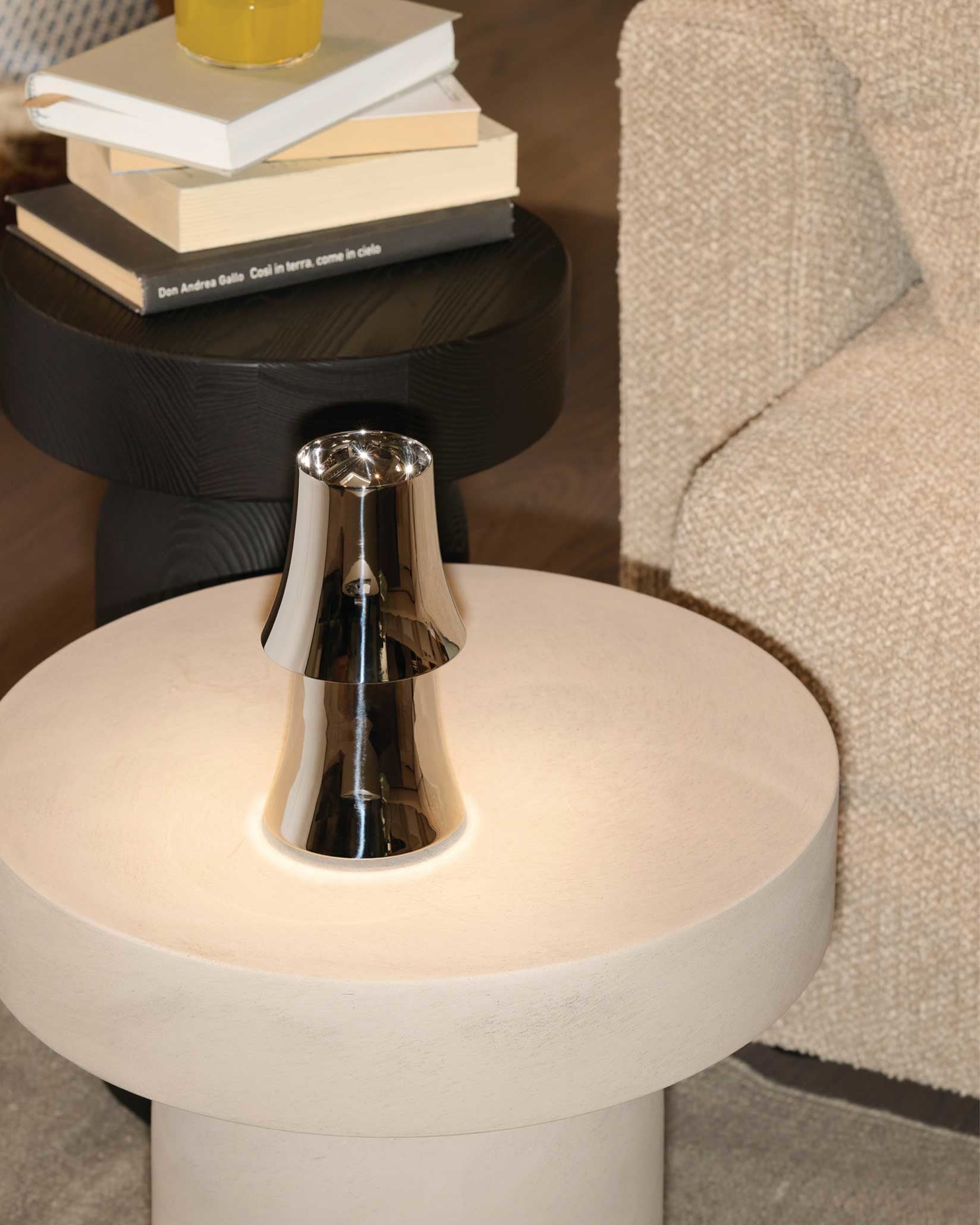 Polished chrome Halesia lamp by Alessi, emitting a soft glow on a light round table next to a beige sofa.