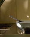 Silver Rocc fragrance diffuser by Alessi, a sleek metallic bowl holding dark incense sticks against a warm, grainy background.
