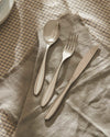 Polished stainless steel table fork by Alessi, alongside a matching spoon and knife on a fringed linen napkin and plaid tablecloth.