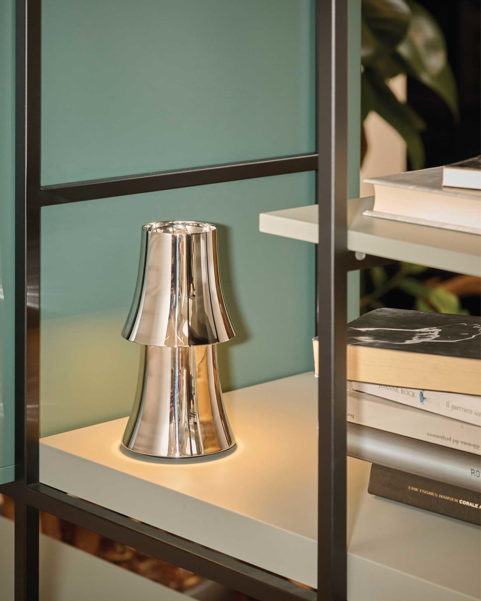 Silver metallic two-tiered lamp by Alessi, illuminated with a warm glow on a white shelf.