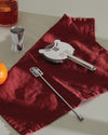 Polished silver-toned mixology accessories by Alessi, including a jigger, strainer, and bar spoon, displayed on a deep red napkin with an orange and a cocktail.