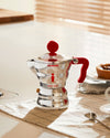 Shiny silver espresso coffee maker by Alessi, with a red handle and knob, featuring A di Alessi and Moka Alessi text with a logo, on a checkered mat.