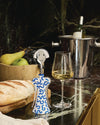 White and blue patterned corkscrew by Alessi with a shiny silver smiling face top, standing upright next to a wine glass.