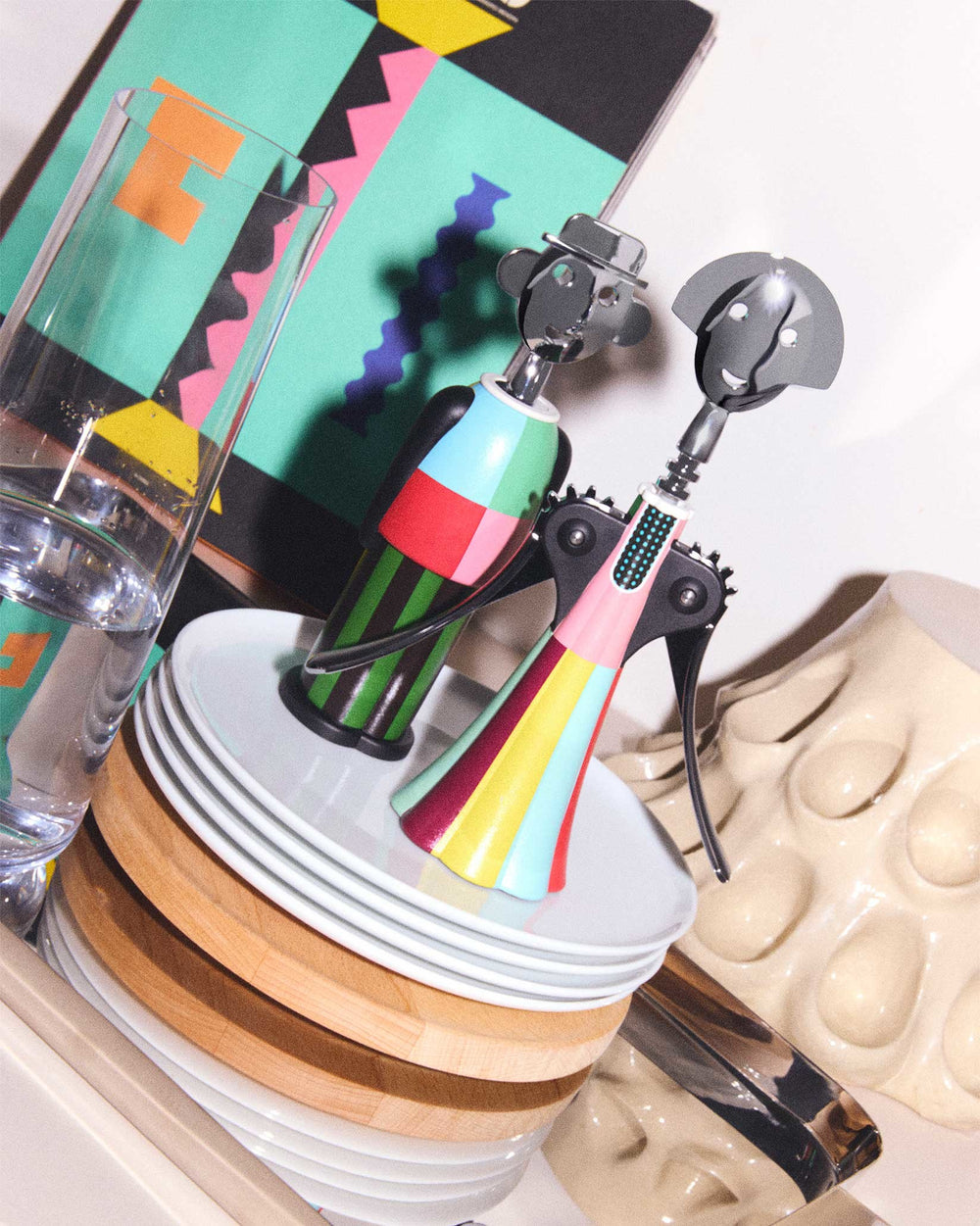 Colorful striped Alessandro M. corkscrew by Alessi, with a hat, displayed alongside a pink figure corkscrew on white plates and wood.