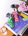 Figurative green and pink patterned corkscrews by Alessi, standing on a magazine with pink flowers and a glass of water.