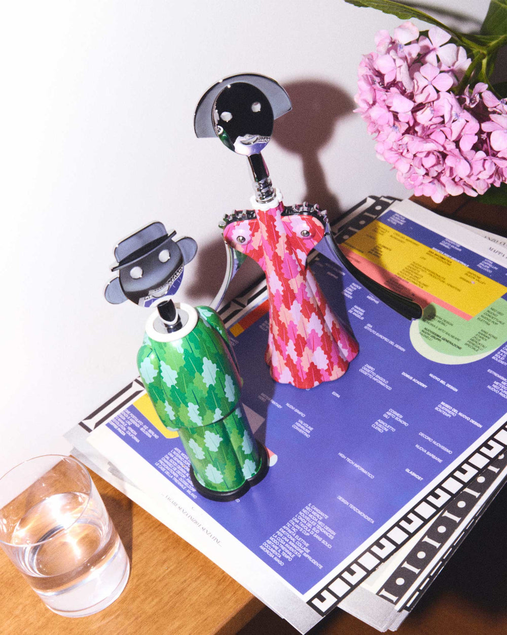 Figurative green and pink patterned corkscrews by Alessi, standing on a magazine with pink flowers and a glass of water.