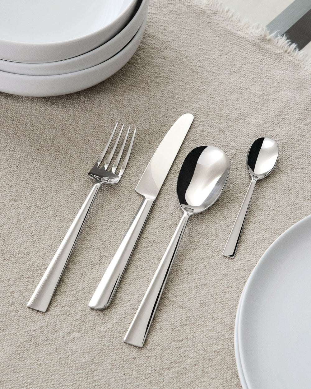 Sleek polished stainless steel flatware set by Alessi, including a fork, knife, and two spoons, on a beige textile surface with white plates.