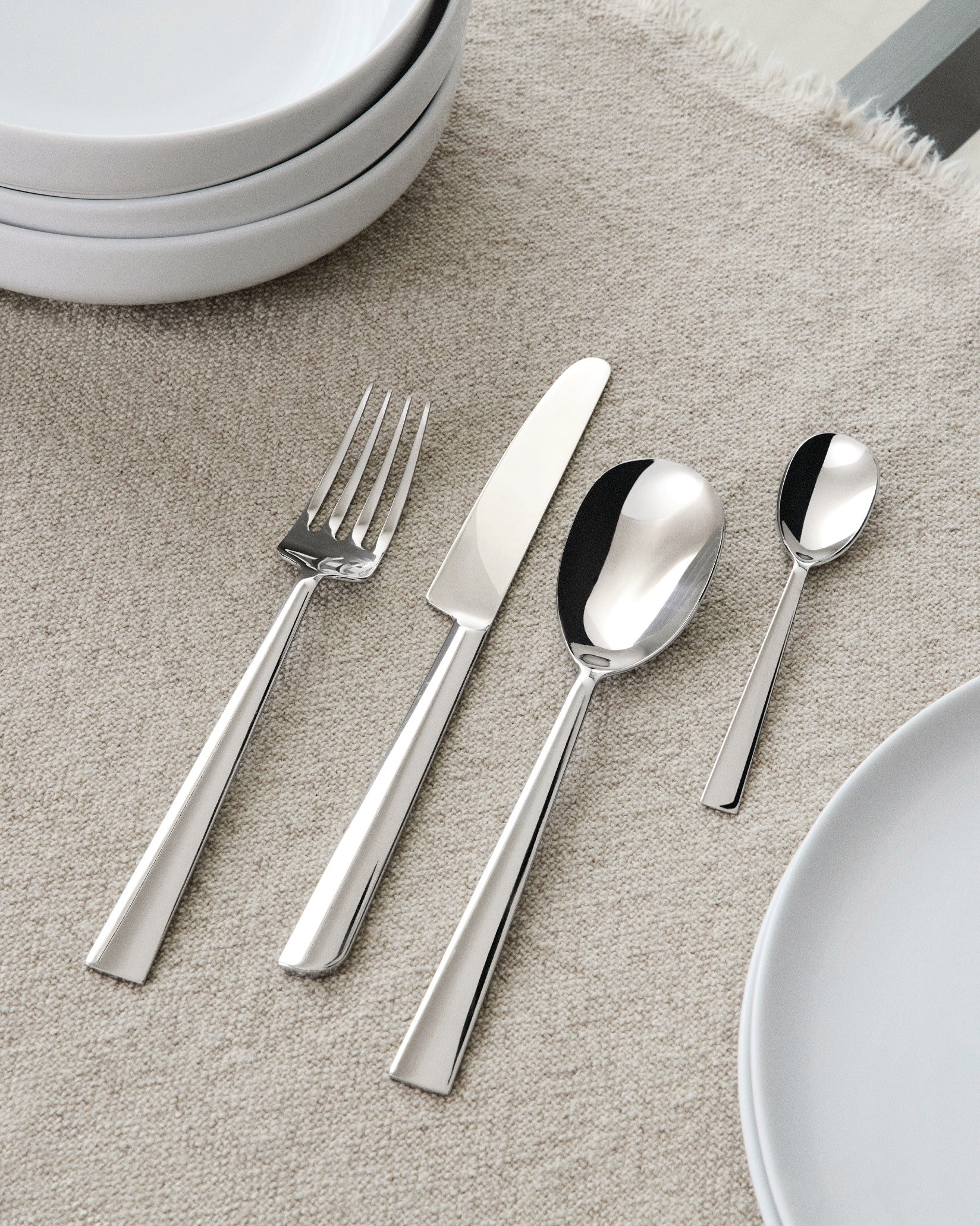 Sleek polished stainless steel flatware set by Alessi, including a fork, knife, and two spoons, on a beige textile surface with white plates.
