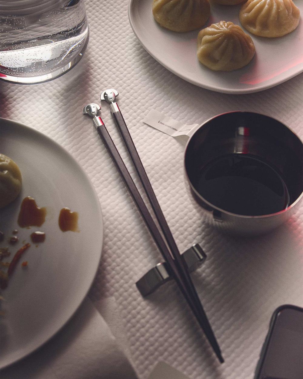 Dark wood chopsticks with silver anthropomorphic heads by Alessi, part of a sushi set with dumplings and soy sauce.