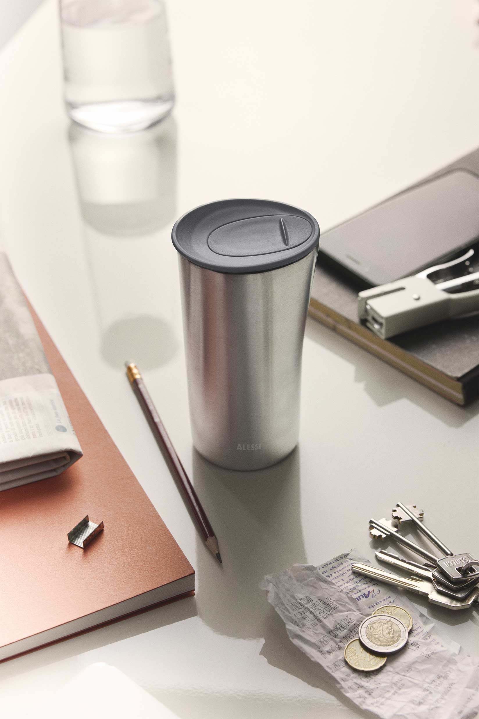 Silver stainless steel thermo insulated tumbler by Alessi with a dark gray lid, on a light desk next to a pencil, notebook, keys, and coins.