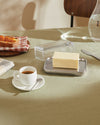 Clear glass and stainless steel butter dish by Alessi holding a block of butter on a light green table.