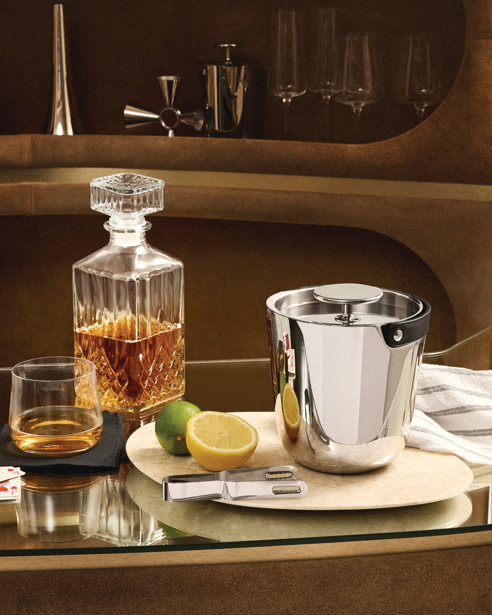 Polished stainless steel ice bucket by Alessi, with a black handle and lid, displayed on a bar with a whiskey decanter, glass, and citrus.