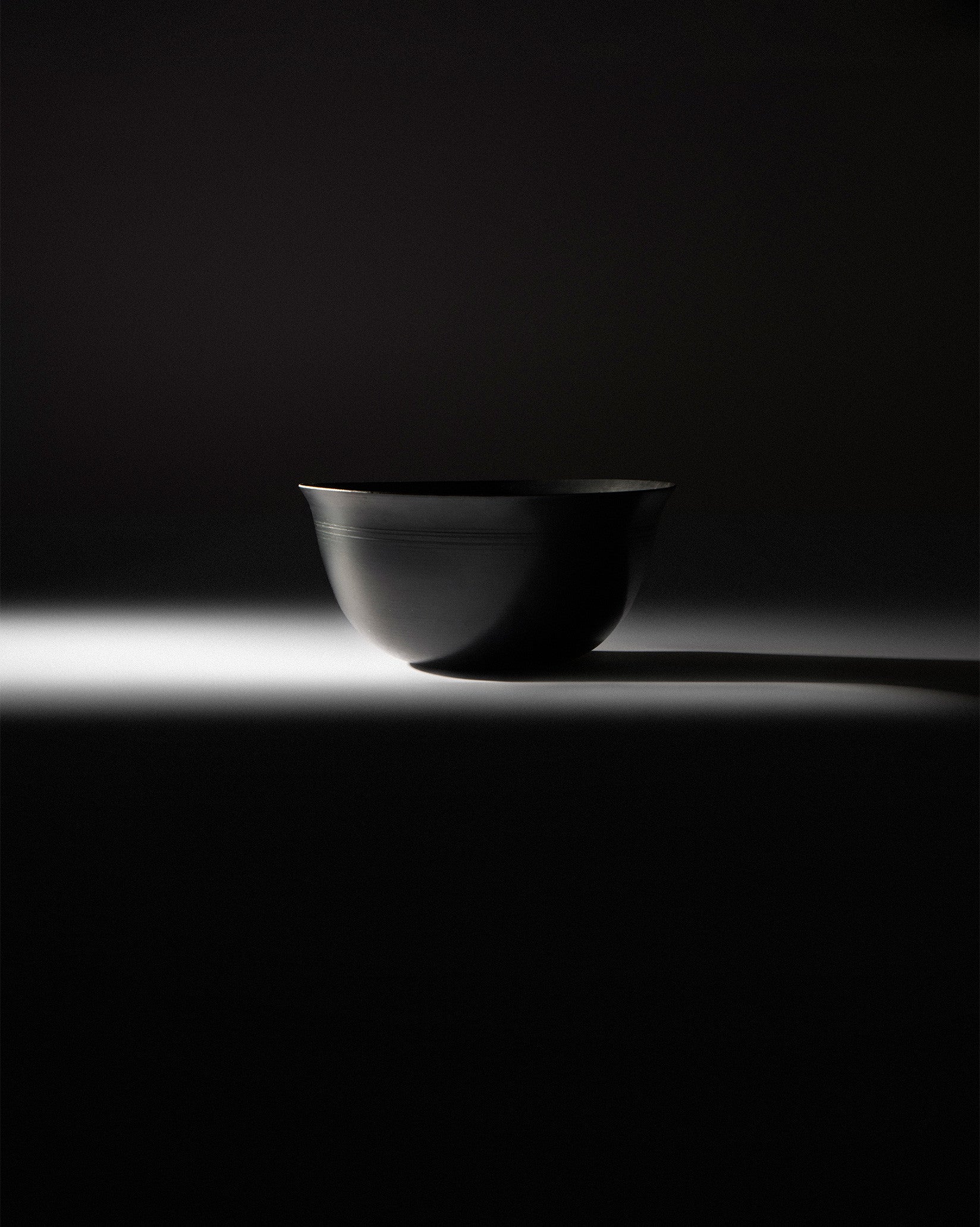 Black metal bowl by Alessi with two etched lines near the rim, dramatically lit from below.