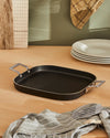 Black rectangular grill pan by Alessi with silver handles and ribbed cooking surface, on a wooden counter with plates and a spatula.