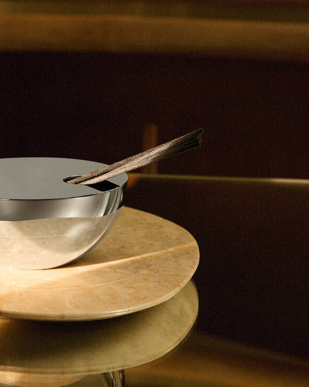 Silver fragrance diffuser by Alessi with dark diffusing sticks on a beige marble surface.
