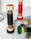 Three multi-colored wood spice grinders by Alessi in a modern bolt-nut design, displayed on a white surface with peppercorns.