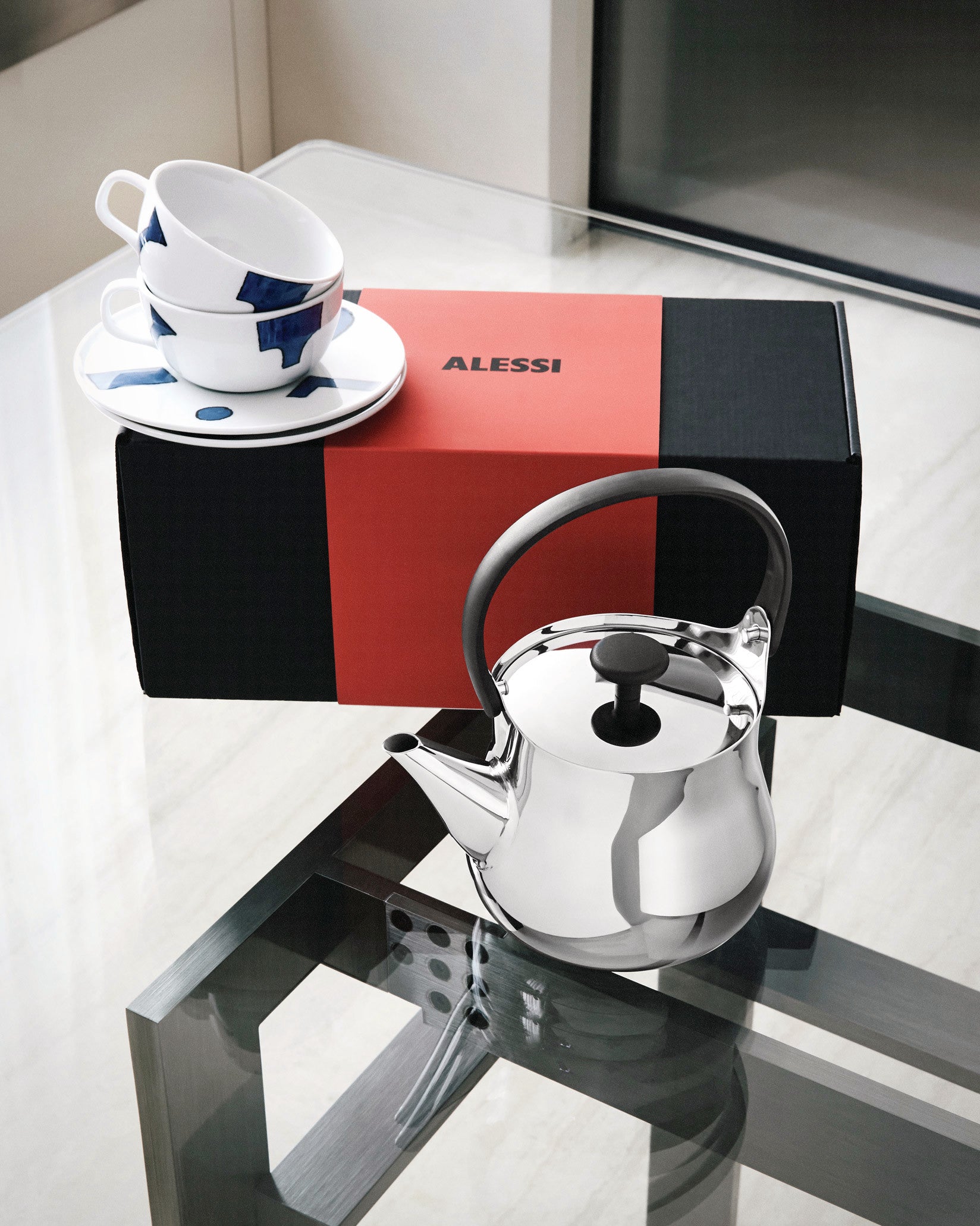 A tea gift set by Alessi, with a shiny silver kettle and two white ceramic cups with blue abstract patterns, displayed next to a red and black box.