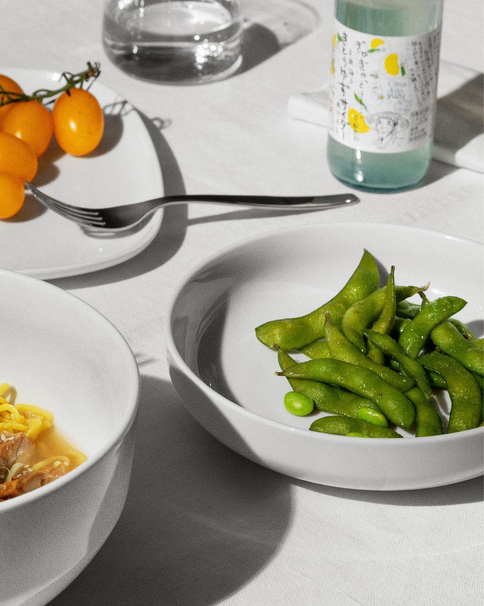 Two white Itsumo soup bowls by Alessi, one with edamame, another with ramen, on a white table.