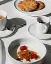 White ceramic tea and coffee set by Alessi with a tea cup, espresso cup, and bowl filled with yogurt and berries.