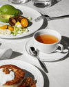 White ceramic teacup and saucer by Alessi, filled with orange tea on a table with breakfast food.