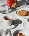 White Itsumo tea cup and saucer by Alessi with amber tea, styled on a white tablecloth with cookies.