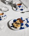 White ceramic table set by Alessi, with a modern blue abstract pattern, displayed on a white tablecloth with drinks and cookies.