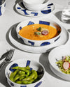 White tableware set with blue geometric patterns by Alessi, displaying soup, edamame, and salad on a white tablecloth.