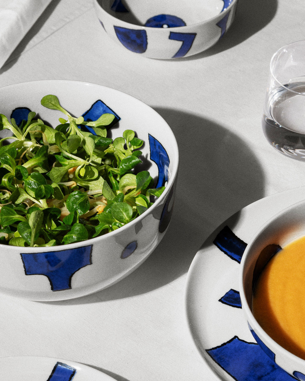 White Yunoki ware table set by Alessi featuring abstract blue patterns, with bowls of green salad and orange soup.