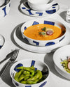 White ceramic table sets by Alessi with blue geometric patterns, holding soup and edamame on a white tablecloth.