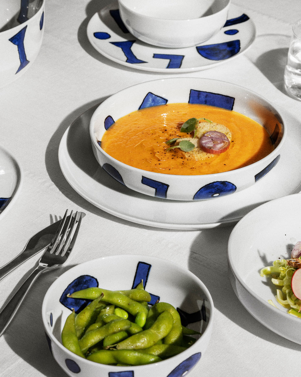 White ceramic table sets by Alessi with blue geometric patterns, holding soup and edamame on a white tablecloth.